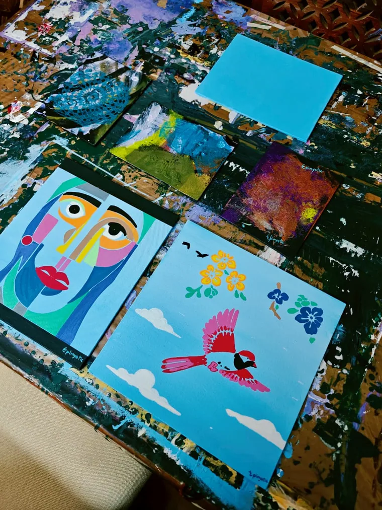 A photograph looking down on a paint-covered wooden studio table holding several small finished canvases: a cubist face portrait, a bird against a blue sky, a blue lace-pattern mandala, a small landscape, a purple and copper textured square, and one blank pale blue canvas waiting.