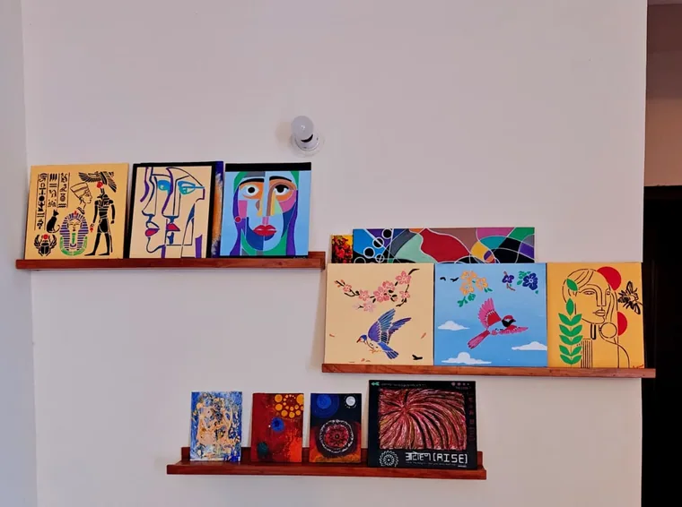 A photograph of a pale wall with three wooden picture-ledges holding many paintings: an Egyptian pantheon panel on yellow, two line-drawn faces, a cubist portrait, a panel of black-outlined colour cells, two bird paintings, a stylised figure with a laurel branch, several small dark abstracts, and the lettered relief painting.