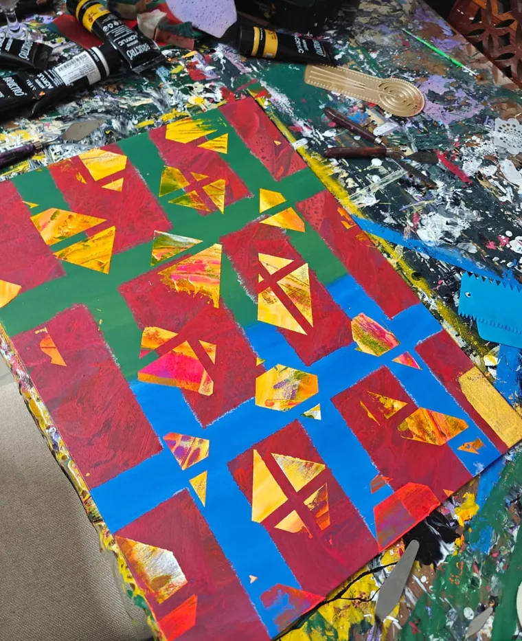 A photograph of a large geometric canvas lying on a heavily paint-covered studio table, its triangles and diamonds in red, green, blue and gold still bright and unshadowed, with tubes of acrylic, palette knives and a toothed scraper around it.