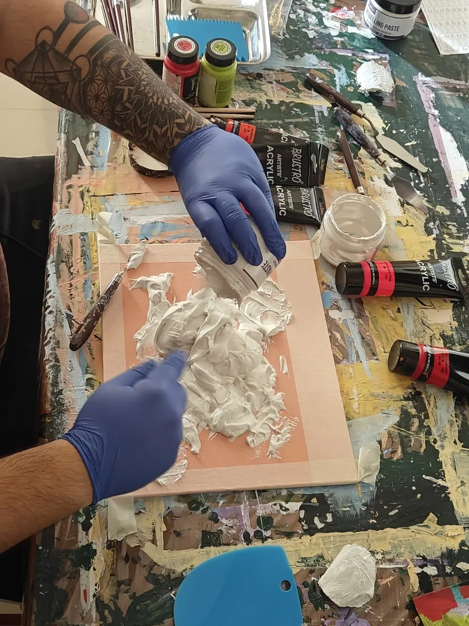A photograph looking down on a pair of gloved hands spreading thick white modelling paste across a taped canvas with a palette knife, surrounded by acrylic tubes, jars, scrapers and painting knives on a paint-covered studio table.