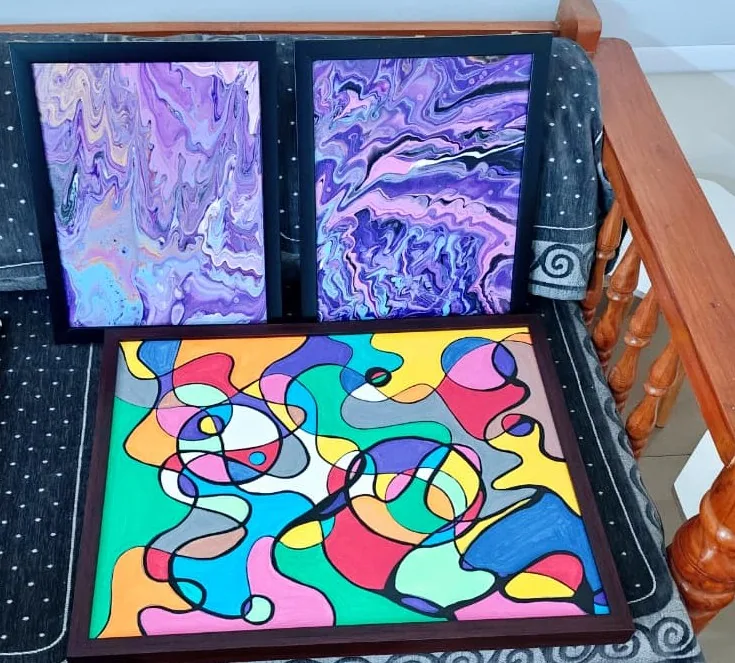 A photograph of three framed paintings resting on a cloth-covered wooden daybed: two upright marbled pours in violet, lilac and white, and a large square painting of black-outlined cells of flat colour in a dark wooden frame.