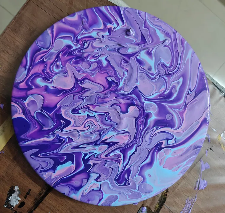 A photograph of a circular canvas of swirling violet, blue, and pink acrylic pour resting on a paint-marked wooden studio table.