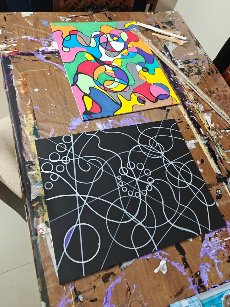 A photograph of two canvases on a paint-splattered wooden studio bench: above, a finished cloisonne painting of black-outlined cells filled with bright flat colour; below, a black canvas bearing only looping white lines and circles, the drawn stage before the colour is added.
