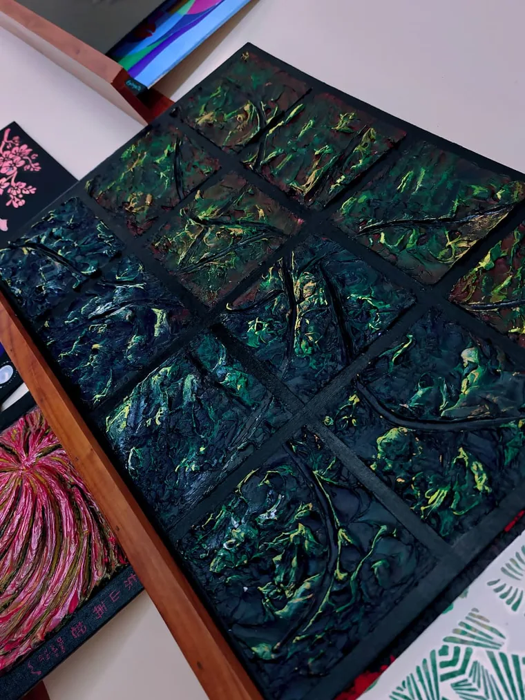 A relief painting divided into nine panes of dark green and black acrylic paste, seen at an angle on a studio table beside other canvases.