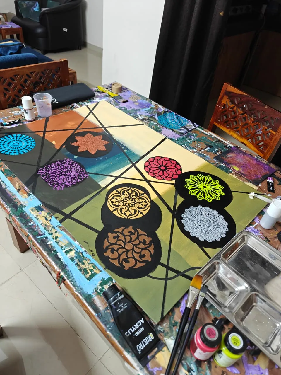 A photograph of a large canvas in progress on a paint-splattered studio table: fields of colour divided by black lines and overlaid with nine circular black discs, each bearing an ornamental mandala in turquoise, copper, violet, crimson, gold, bronze, neon green or silver, with paint tubes, bottles, palette and brushes around it.