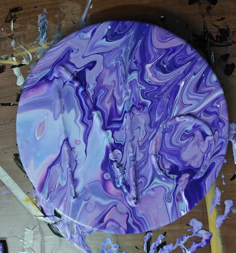 A photograph of a circular canvas of glossy violet, lilac, and white acrylic pour, still wet, on a paint-splattered wooden studio surface.