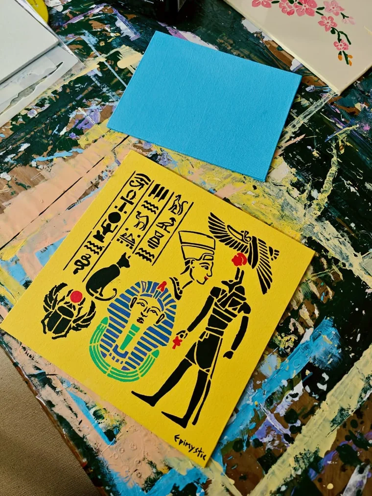 A flat graphic painting on a bright yellow ground: bands of black hieroglyphs, a scarab beetle with a red sun disc, a seated black cat, the blue-and-gold mask of Tutankhamun, Nefertiti in profile with her crown, and a winged figure holding an ankh.