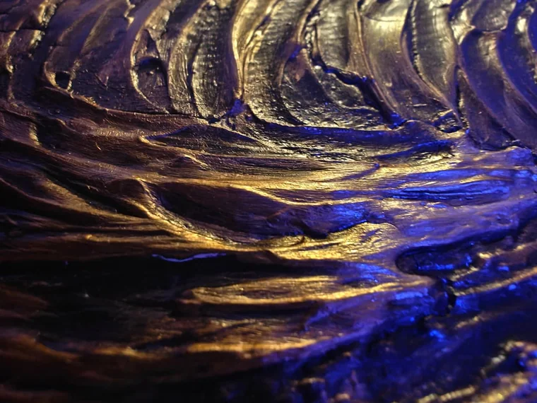 Extreme close-up of a sculptural relief painting of a swirling, vortex-like surface, lit warmly so the raised ridges read in bronze, gold, and violet against deep shadow.