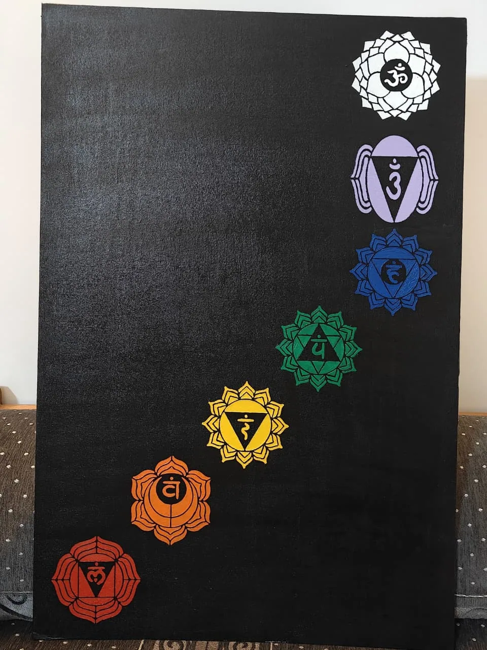 A photograph of a tall black canvas painted with the seven chakra symbols in an ascending diagonal, from the red root wheel at the base to the white crown wheel with the Aum at the top, leaning in a room.
