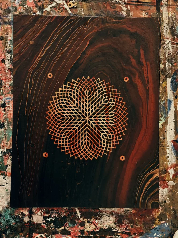 A lattice of fine gold lines crossing into an eight-petalled star at the centre of a dark red-brown panel painted with wood grain, four gold dots around it, on a paint-spattered studio floor.