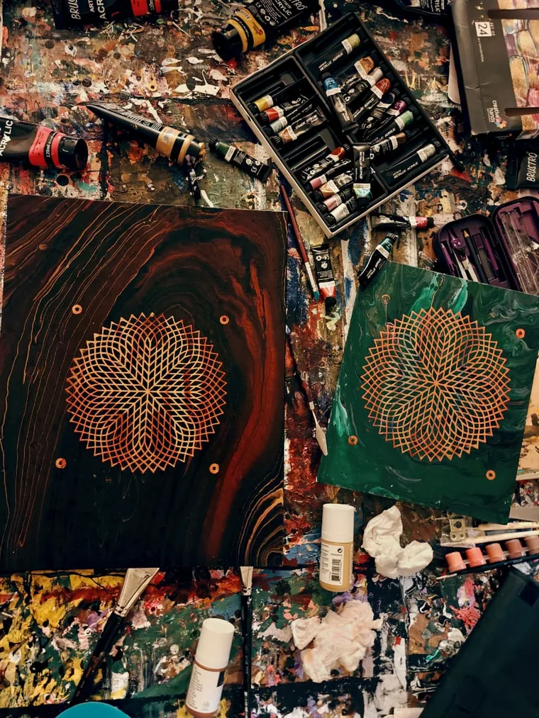 Two panels on a paint-covered workbench, each with a gold line-lattice star, one on dark wood grain and one on marbled green, surrounded by paint tubes, brushes and rags.