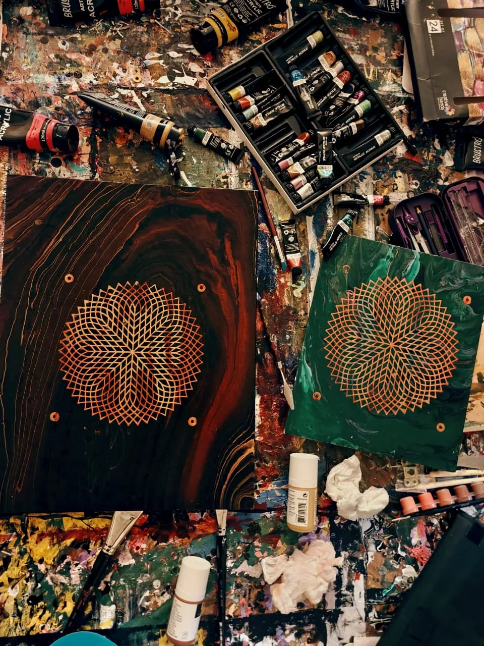 Two panels on a paint-covered workbench, each with a gold line-lattice star, one on dark wood grain and one on marbled green, surrounded by paint tubes, brushes and rags.