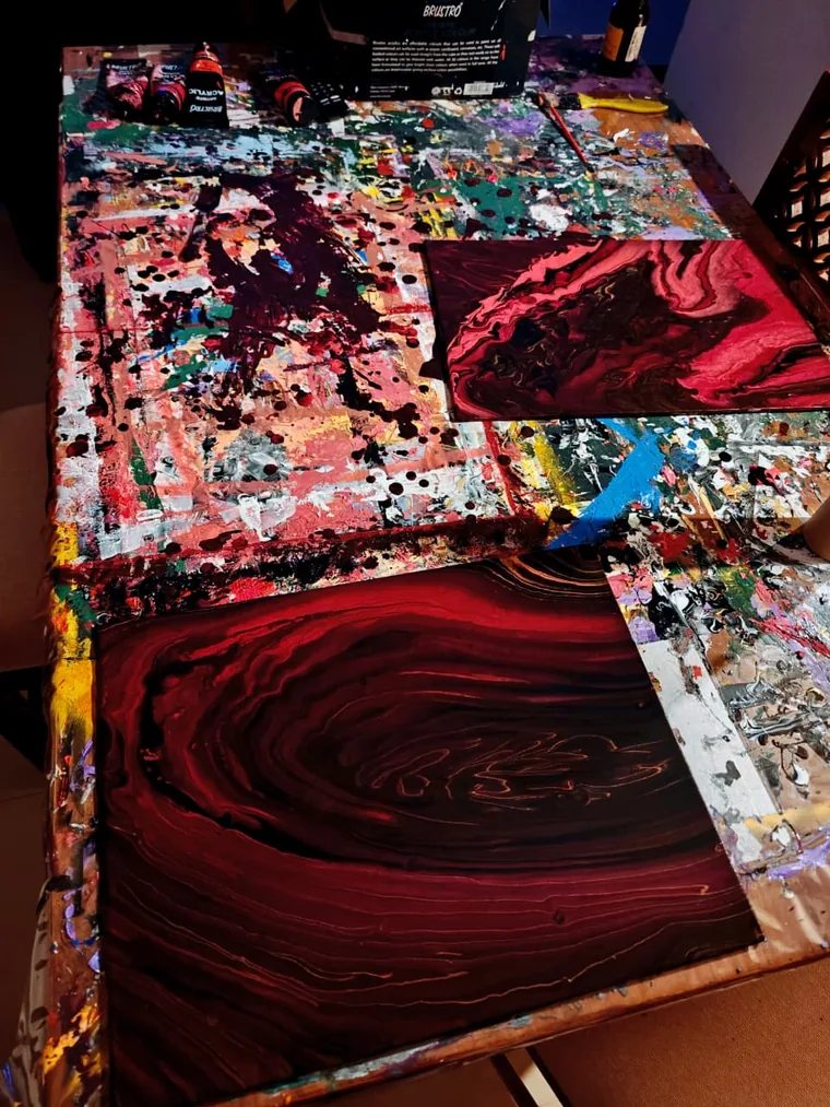A photograph looking down a heavily paint-spattered studio table at two freshly poured canvases lying wet: a large one in deep oxblood with a wide whorl through it, and a smaller one in crimson and pink, with tubes of acrylic and a paint-caked palette around them.